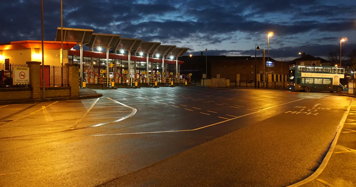 An empty bus station forecourt lit by sodium lights at dusk, with a single double-decker bus parked at a stand, Ossett, West Yorkshire, England.