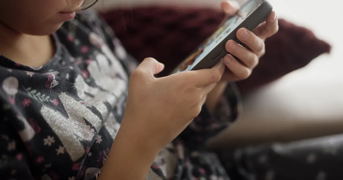 A child sits on a sofa at home, holding a smartphone with both hands, face turned down toward the screen.