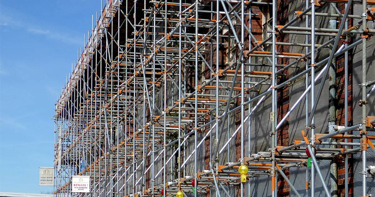 A multi-storey scaffold structure wrapped in debris netting against the brick wall of a UK building under renovation.