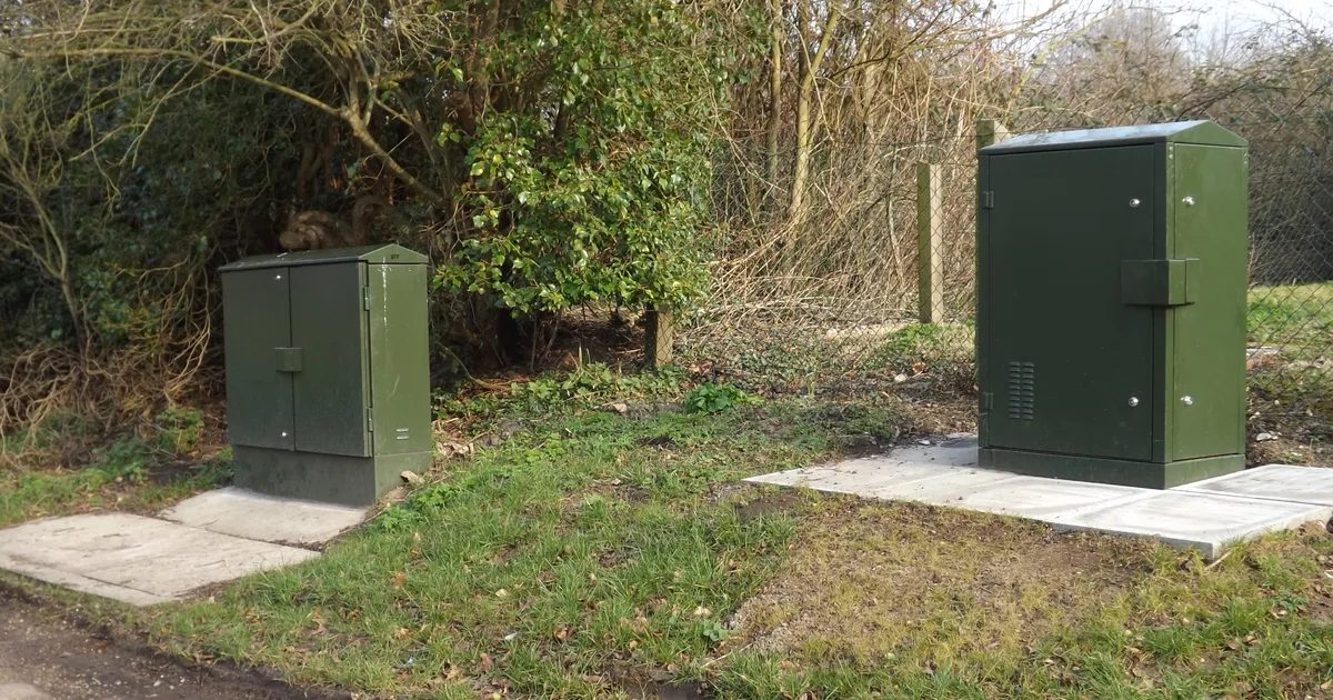 Two green British broadband fibre cabinets on a grass verge beside a rural road, surrounded by hedges and bare trees.