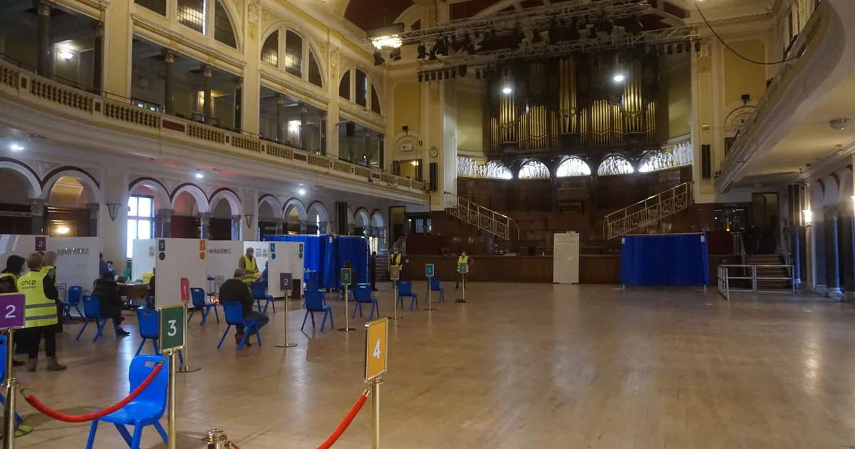 A mass Covid-19 vaccination centre set up inside a grand civic hall, with numbered booths, blue screens and volunteers in hi-vis vests on the hall floor.