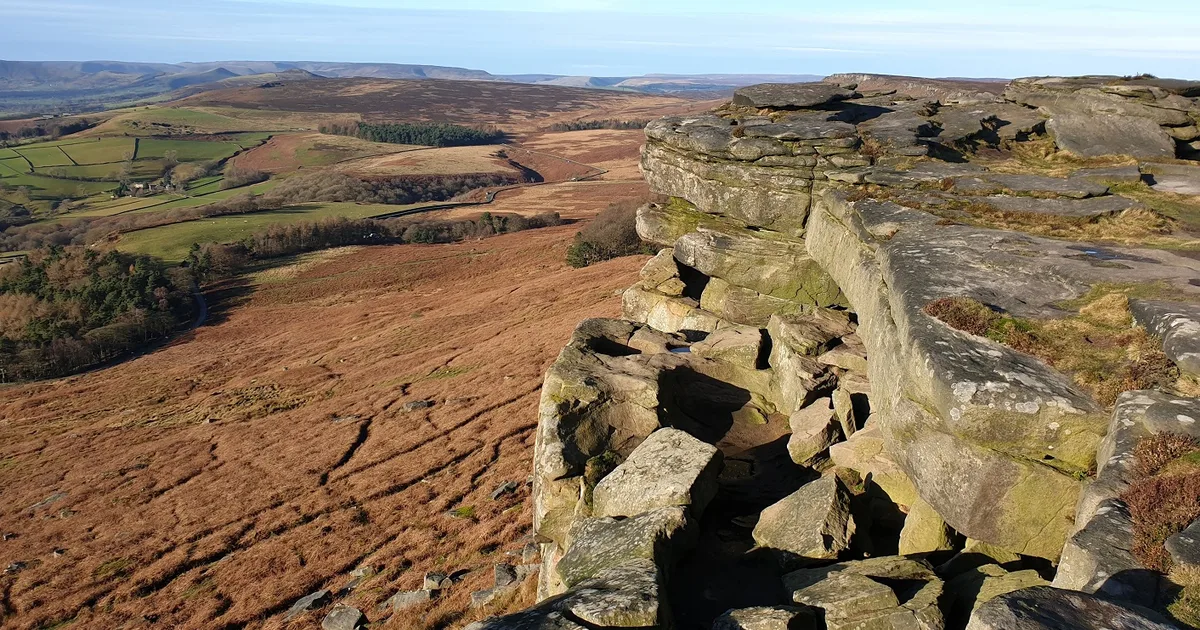 Stanage Edge, a gritstone escarpment in the Peak District National Park, England, seen from above with moorland and farmland stretching into the distance.