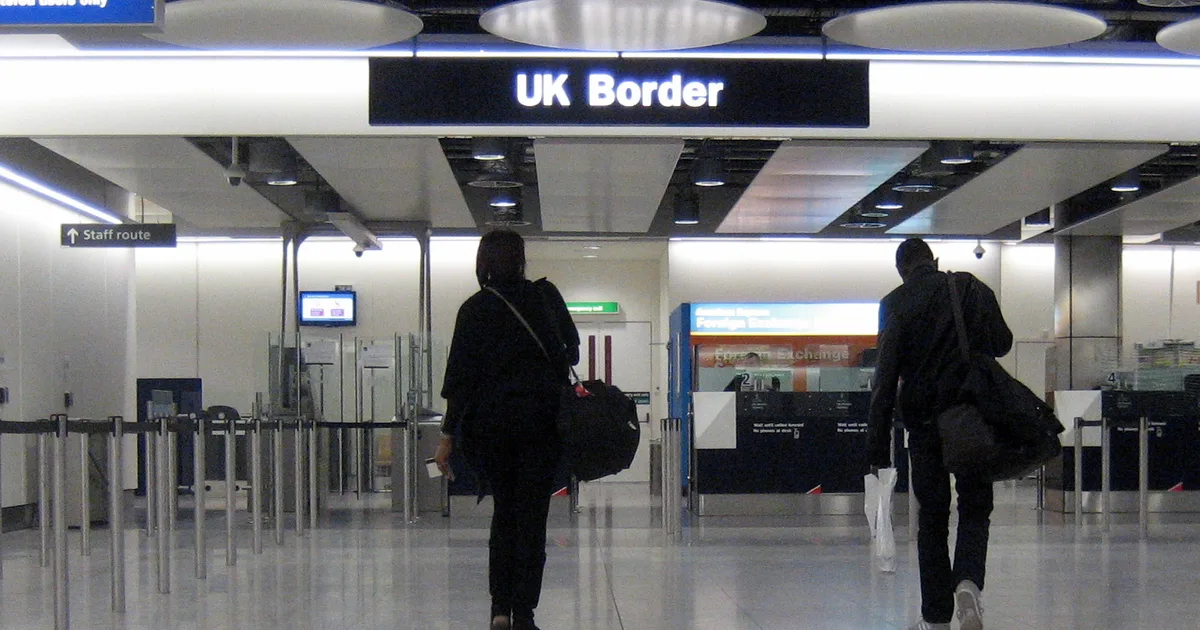 Two travellers walk toward the UK Border sign and passport gates at Heathrow Airport.