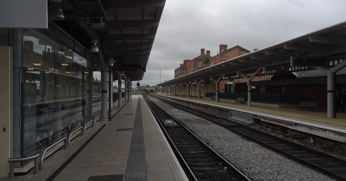An empty platform at Derby railway station, with a canopy roof, railway tracks and Victorian station buildings visible under a grey sky.