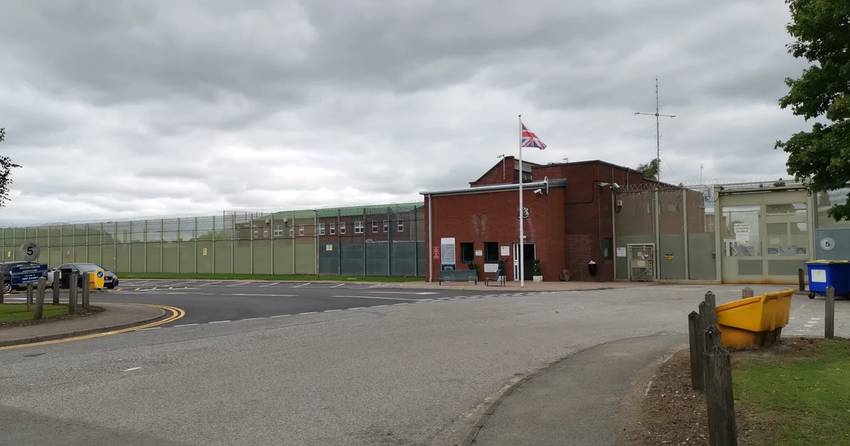 The gatehouse and perimeter fence at the entrance to HM Prison Ranby, a red-brick building with a Union flag behind a high mesh fence.