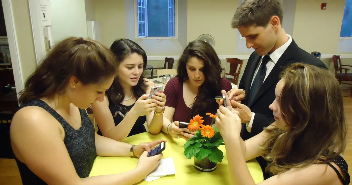 Five young people gathered around a table, each looking down at a smartphone in their hands.