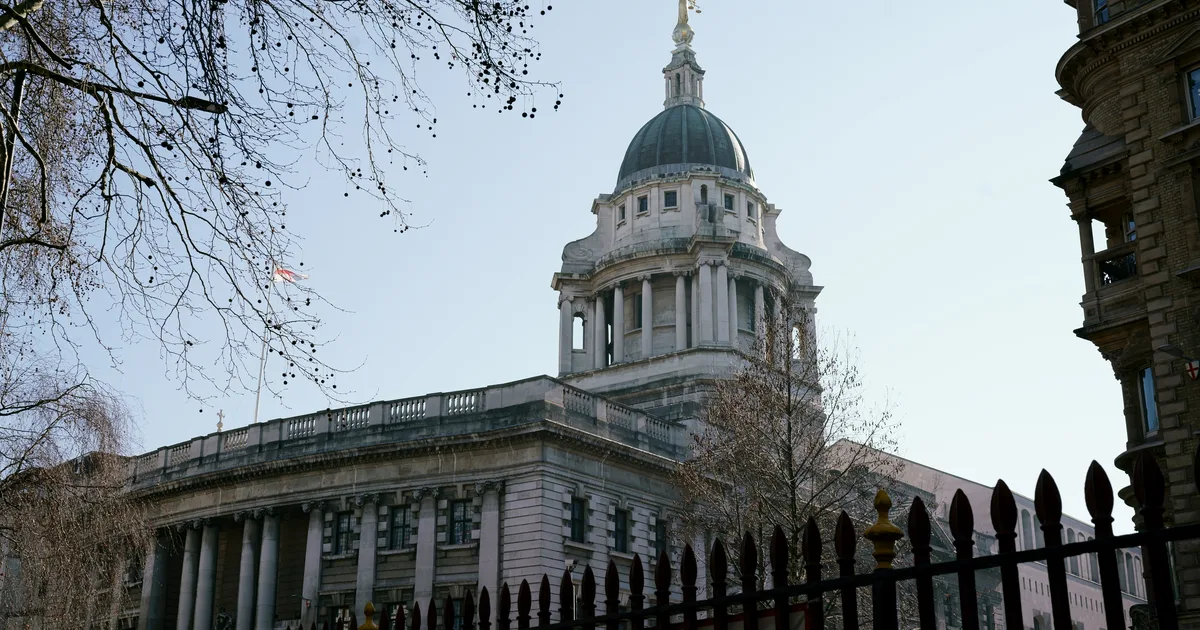 The dome of the Old Bailey, Central Criminal Court, topped by the gilded figure of Lady Justice, seen through bare branches and iron railings.