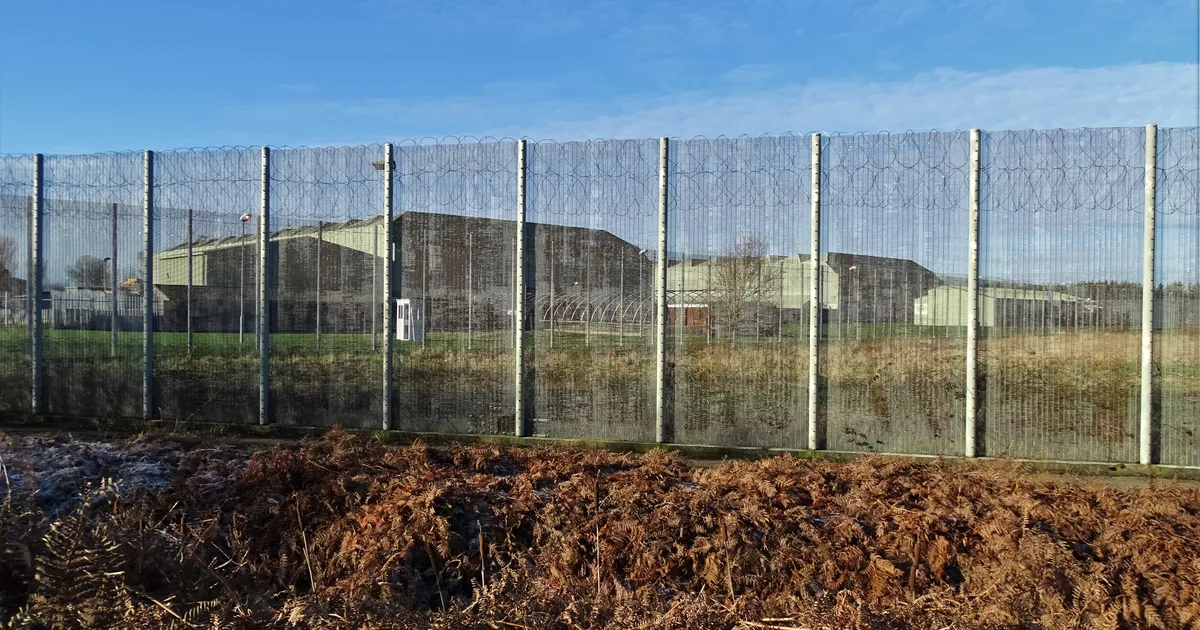 The perimeter fence at HMP Lindholme, topped with razor wire, with the prison's repurposed RAF-era buildings visible behind it.