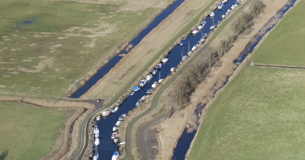Aerial view of moored boats along a narrow Norfolk Broads waterway, running between grazing marshes.