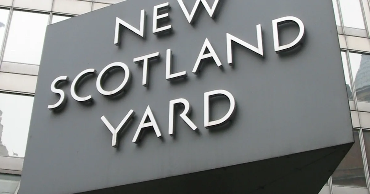 The New Scotland Yard sign, a grey triangular block lettered in white, outside a glass office building.