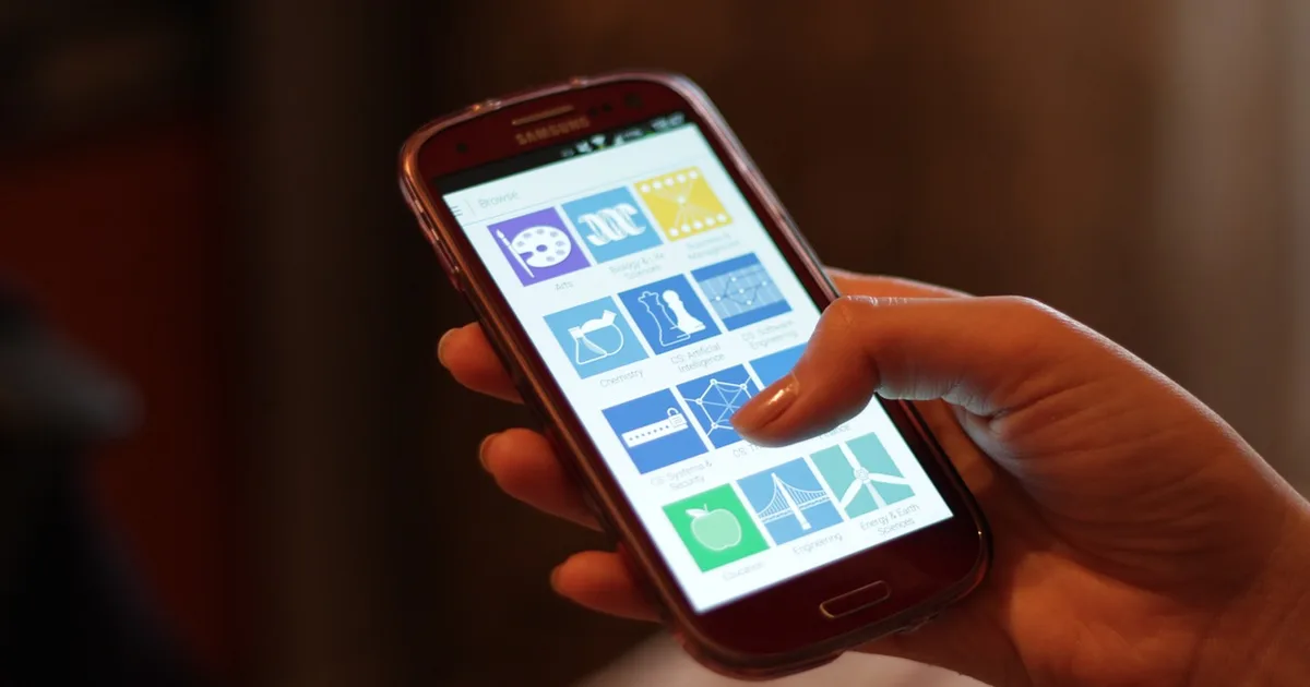 A hand holding a smartphone, thumb on the home screen, showing a grid of app icons.