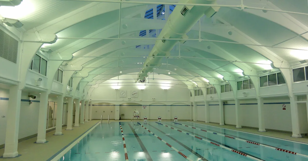 An empty indoor public swimming pool with lane ropes, a vaulted white roof and skylights, at Dulwich Leisure Centre, London.