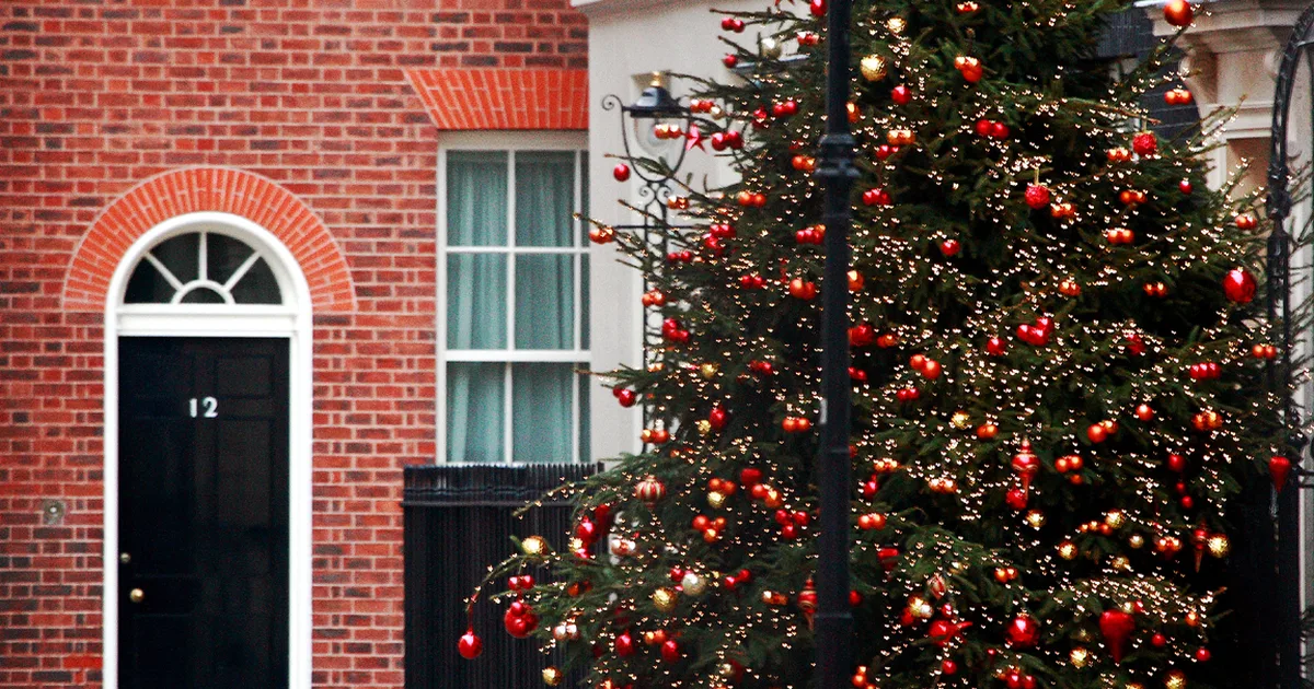 A Christmas tree decorated with red and gold baubles outside a brick townhouse on Downing Street, London, with a black front door numbered 12 visible to the left.