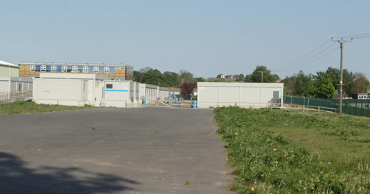Temporary classroom blocks on a tarmac yard at a secondary school, with the main school building and fields behind.