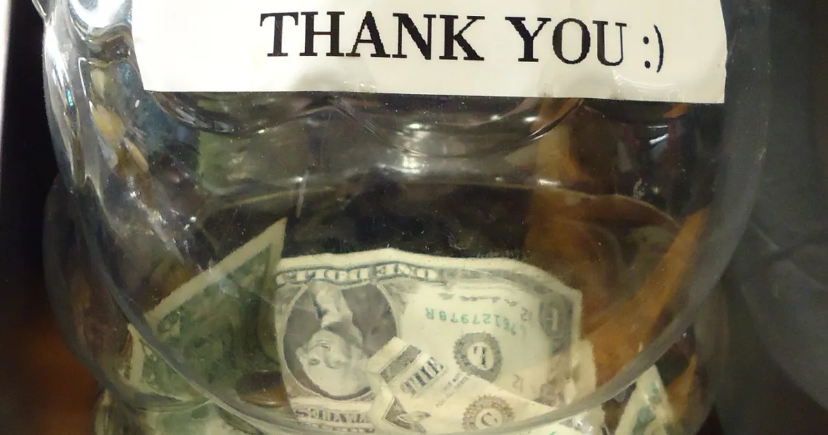 A tip jar on a counter, labelled THANK YOU, holding dollar bills and coins.